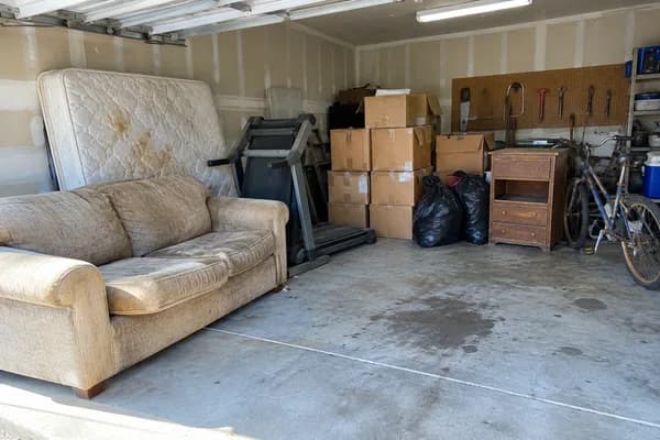 A garage with an old couch, a stained mattress, a treadmill, boxes and trash bags